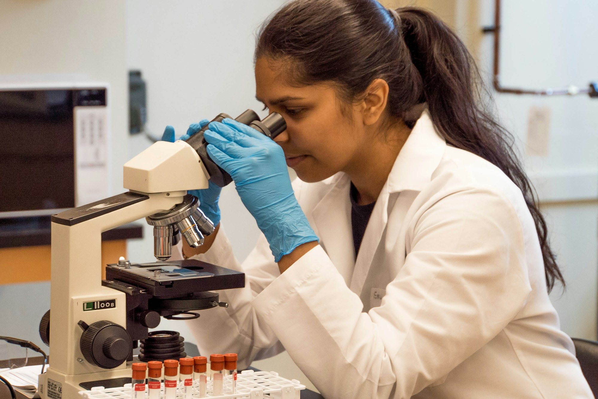photo-1562789233-495f52b583dd A woman looks through a microscope as she examines samples in a laboratory.