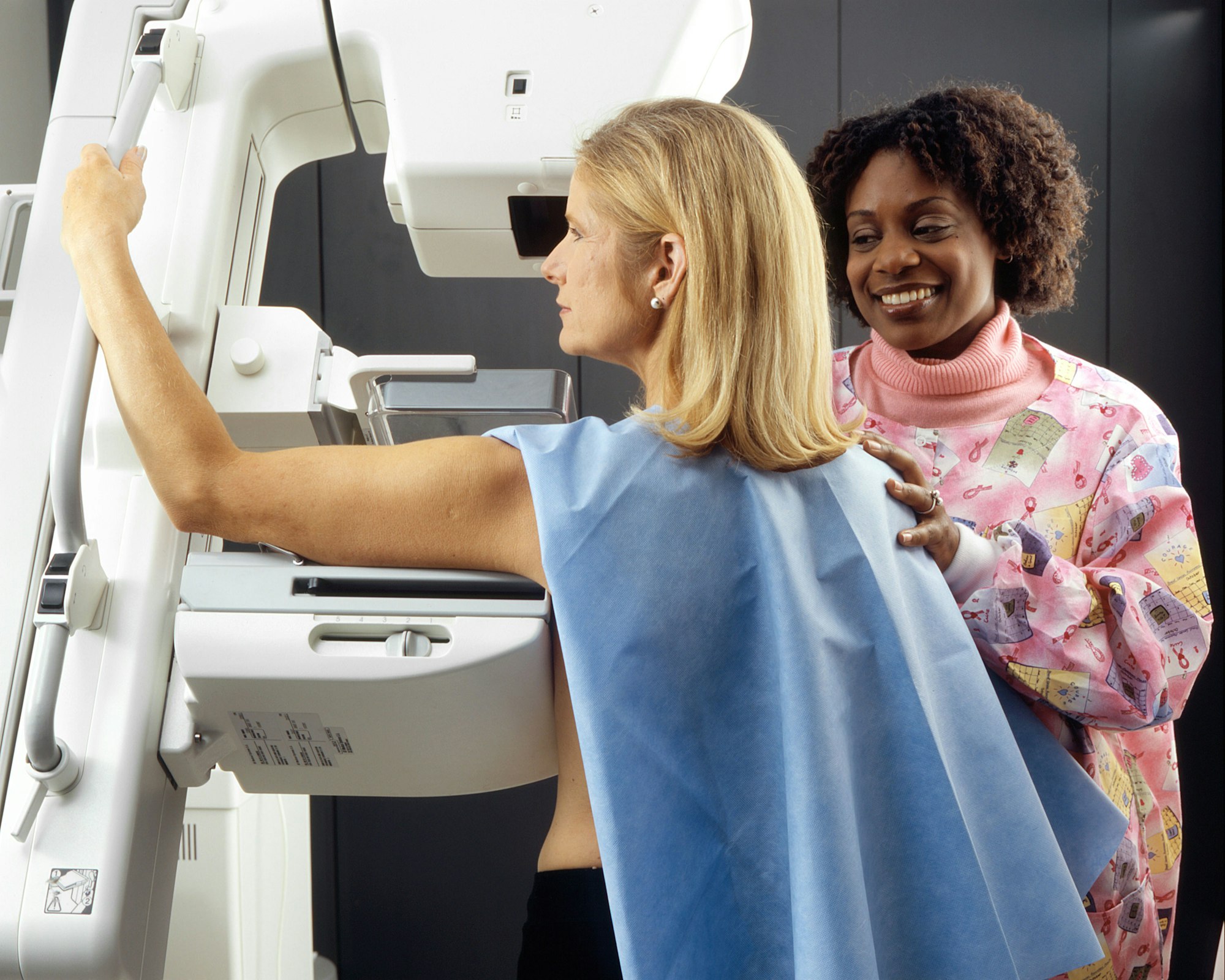 photo-1579154341098-e4e158cc7f55 A smiling technician positions a woman at an imaging machine for a mammogram.