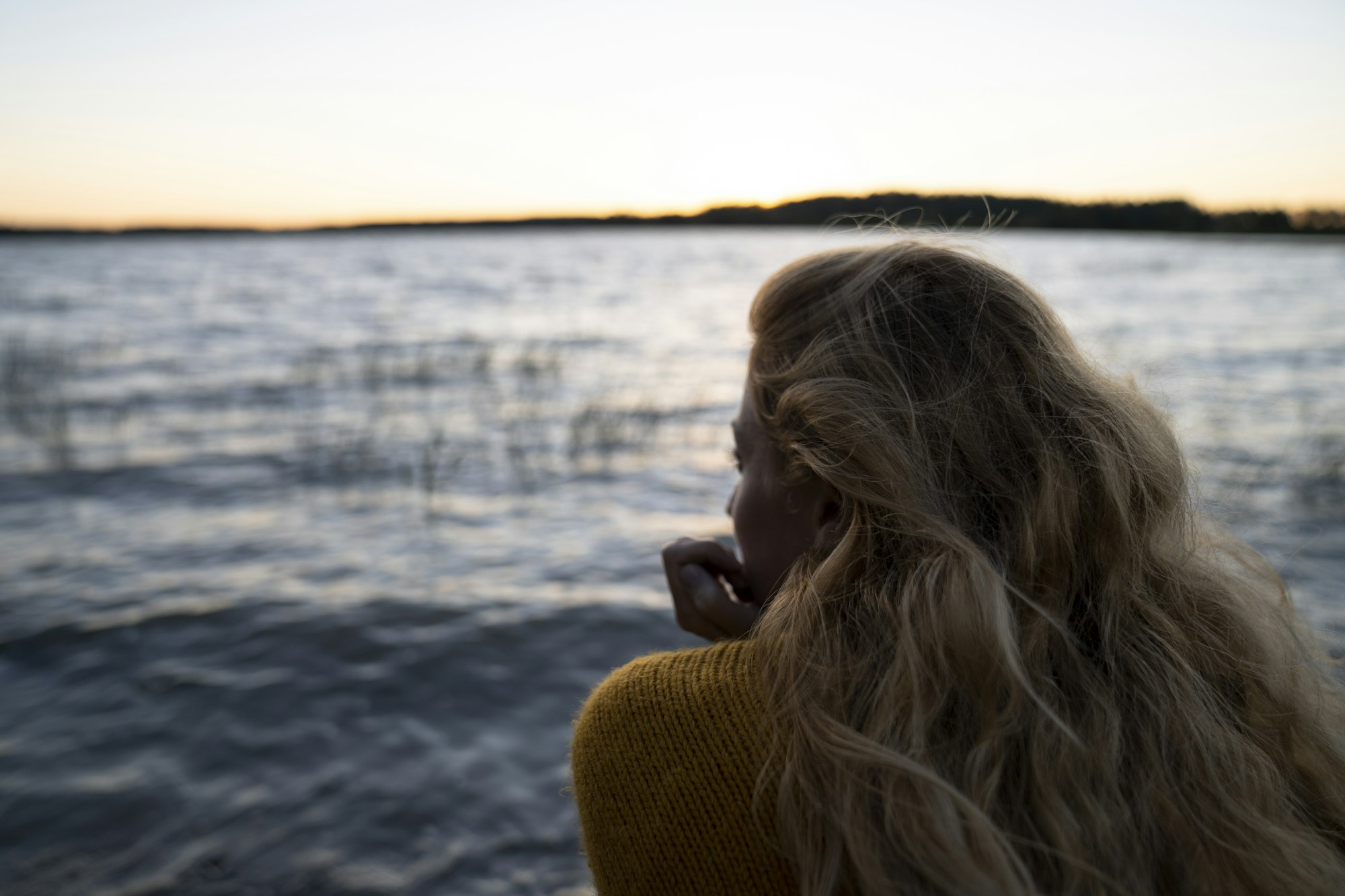 photo-1623307201324-594f7d7e804d A woman with long blonde hair looks out over a lake as the summer sun sets behind the forest in Finland.