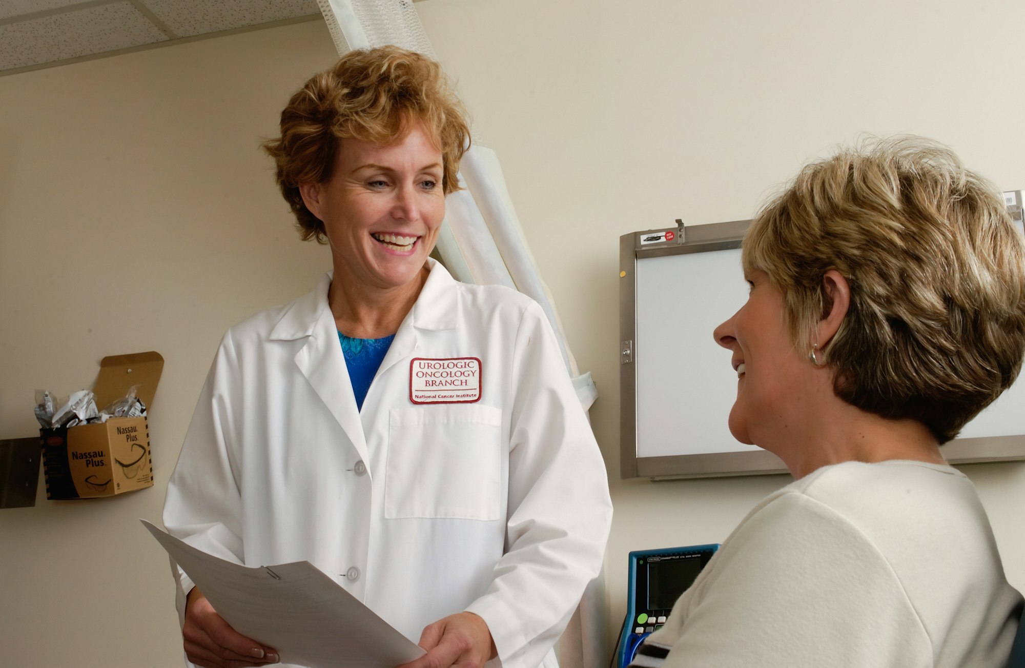 photo-1631217868264-e5b90bb7e133 A smiling doctor in a white coat talks with a patient in a clinic.