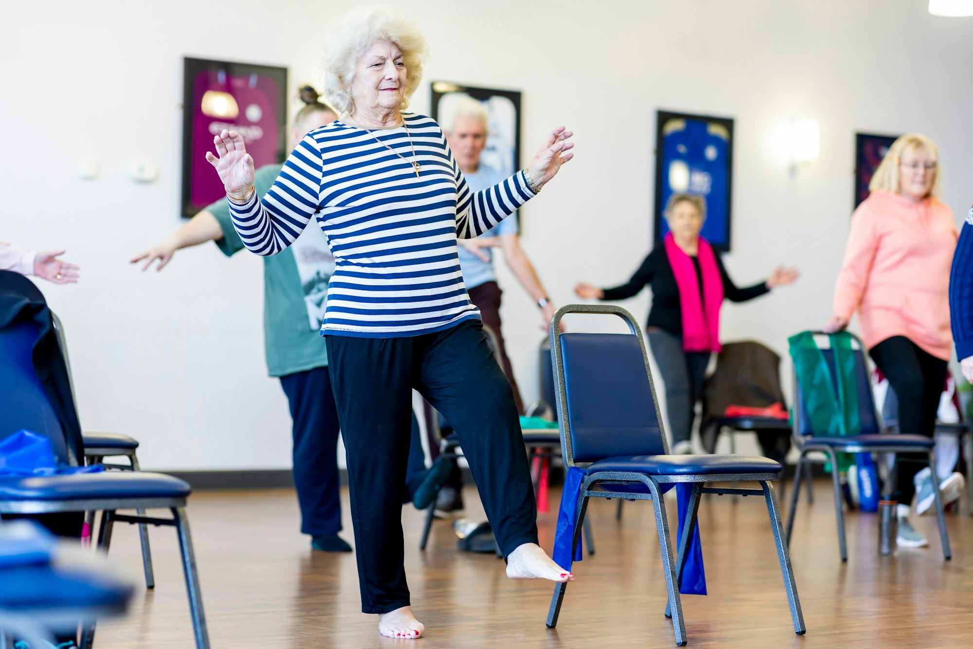 photo-1702648158754-484755fadf79 A group of older women take part in an exercise class, dancing together in a room.