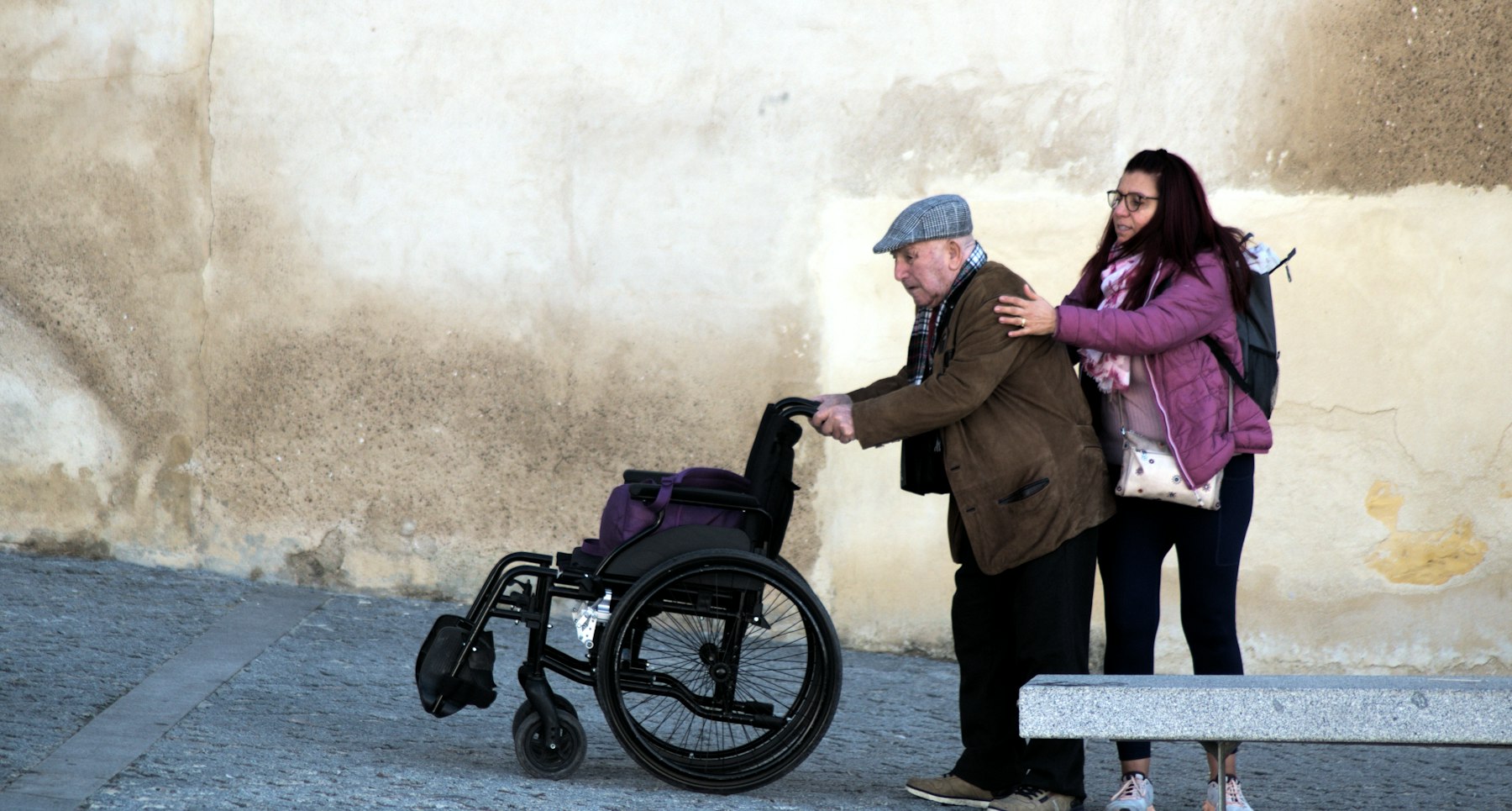 photo-1764006145420-df3006edf060 A young woman helps an older man who is trying to walk with his wheelchair.