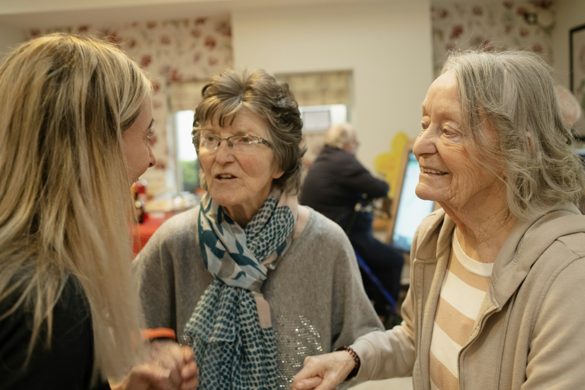 photo-1773227059745-e4ed390faba4 Three older women talk and smile together in a room.